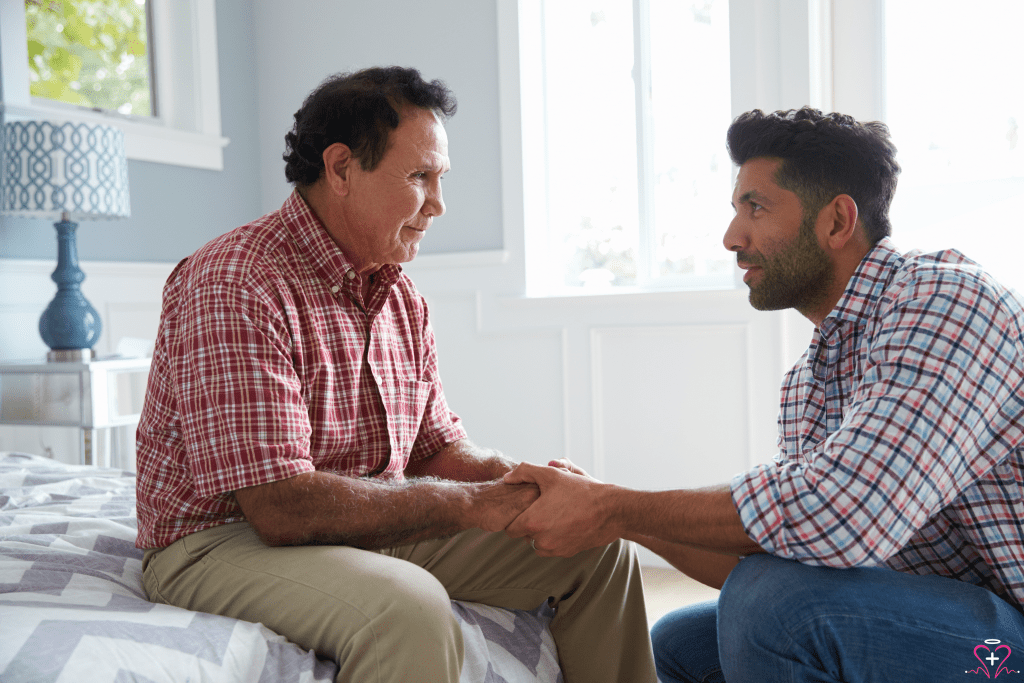 A younger man comforting an older man who is sitting on a bed, holding his hands. Bereavement Services