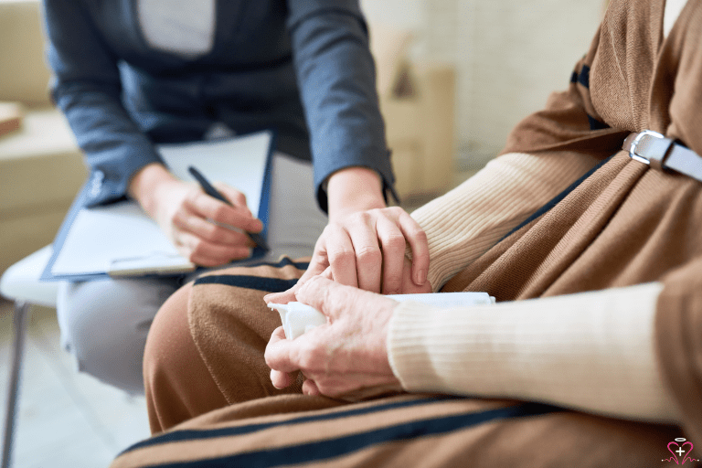 Spiritual Support - Close-up of a caregiver holding the hand of an elderly person while taking notes on a clipboard.