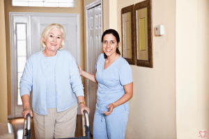 The Importance of Home Health Aides - An elderly woman using a walker with the assistance of a home health aide in a residential hallway.