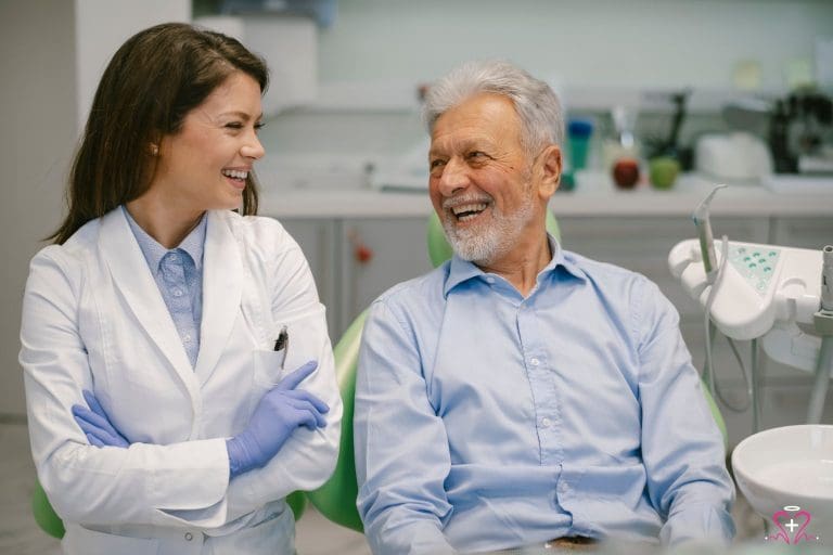 A senior man smiles while sitting next to a friendly dentist during his dental appointment, symbolizing affordable dental care.