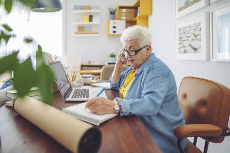 Older adult speaking by phone while taking notes beside a laptop