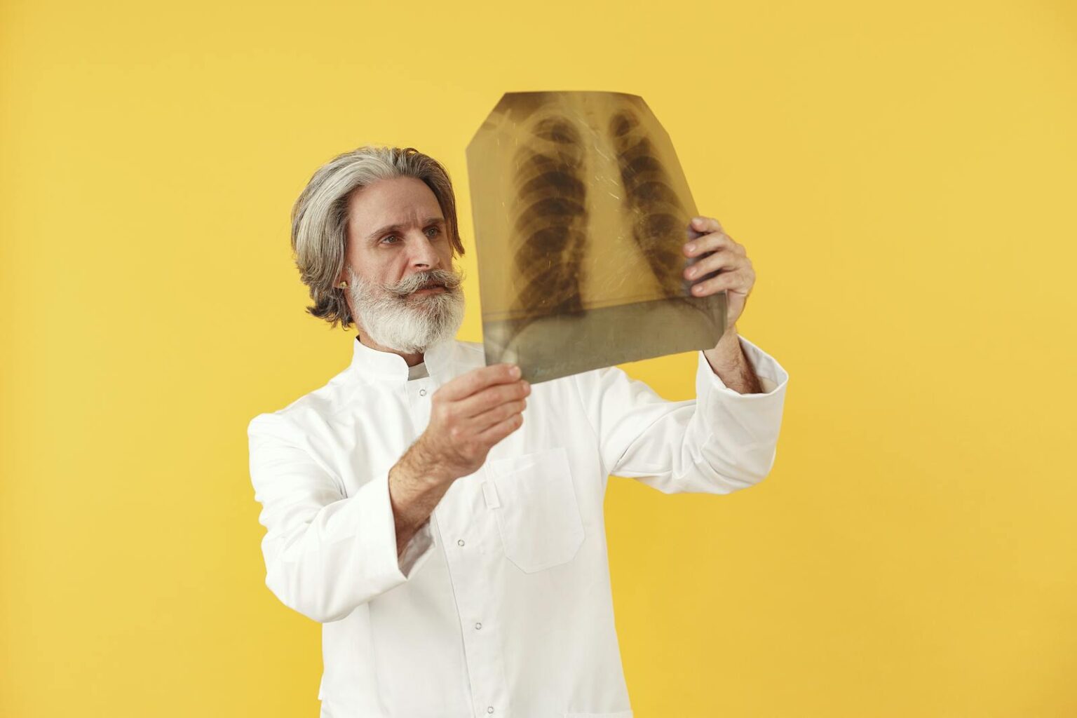 Senior male doctor holding and examining a chest x-ray, posing against a bright yellow studio background.