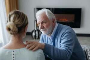 An elderly man comforts a woman indoors near a cozy fireplace, expressing emotional support.
