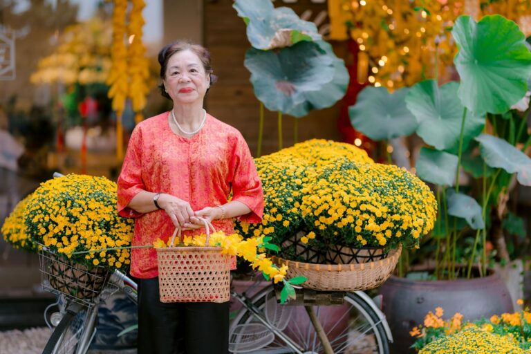 Elderly woman poses with bicycle overflowing with vibrant yellow chrysanthemums outdoors.