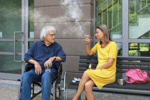 Senior man in wheelchair and woman in yellow dress smoking outside urban building.